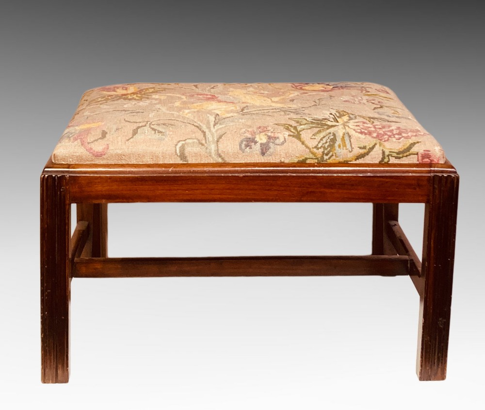 An antique mahogany rectangular dressing stool featuring fluted legs, an H-stretcher base, and a padded floral tapestry seat.