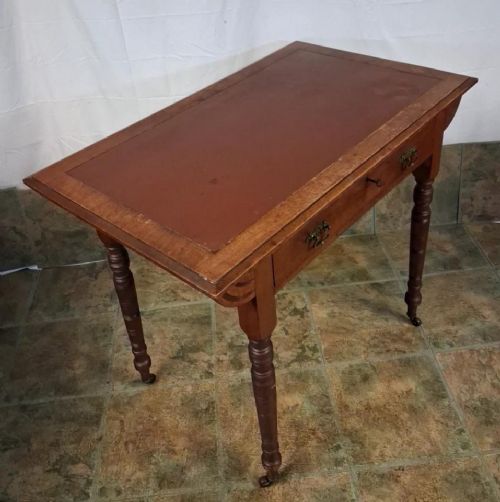 fig 1. A small Victorian mahogany writing table featuring a reddish-brown leather inset top, a single drawer with brass drop handles, turned legs, and brass castors.