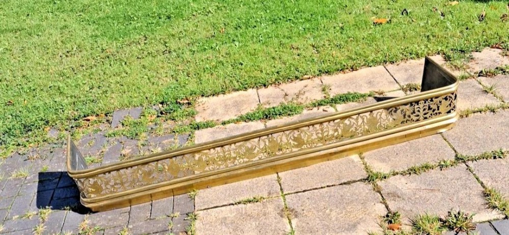 An antique brass fire fender featuring intricate pierced fretwork depicting urns and mythical winged creatures along the front rail.