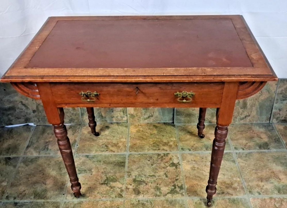 A small Victorian mahogany writing table featuring a reddish-brown leather inset top, a single drawer with brass drop handles, turned legs, and brass castors.