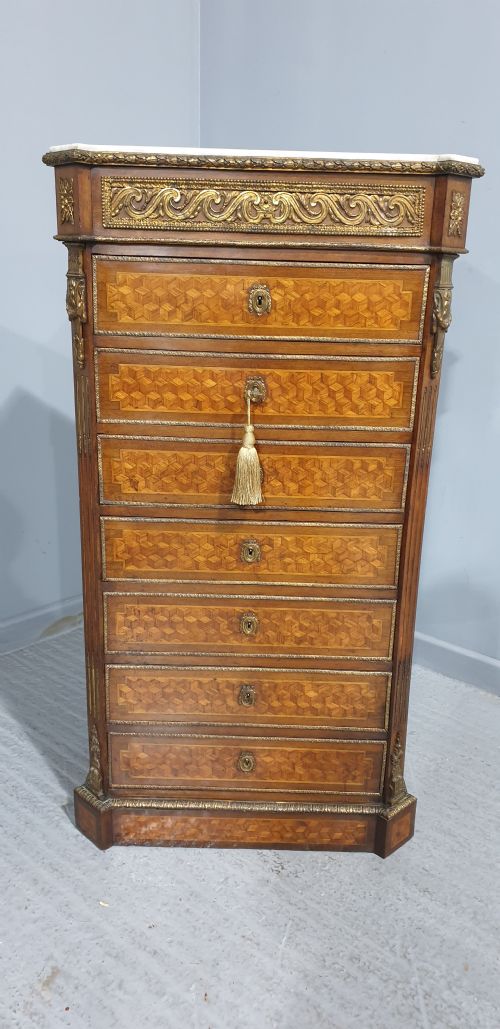 fig 1. A tall French seven-drawer semainier chest with geometric wood parquetry, gold-toned ornamental mounts, and a white marble top.