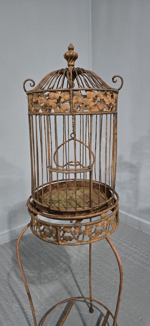 fig 1. A Victorian cast iron birdcage featuring a domed roof and decorative filigree band, supported on a tall slender metal floor stand with an oxidised rust finish.
