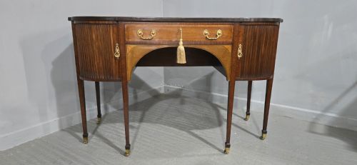 fig 1. A George III style inlaid mahogany sideboard featuring a central drawer above an arched aperture, flanked by vertical fluted cupboards on tapered legs with brass castors.
