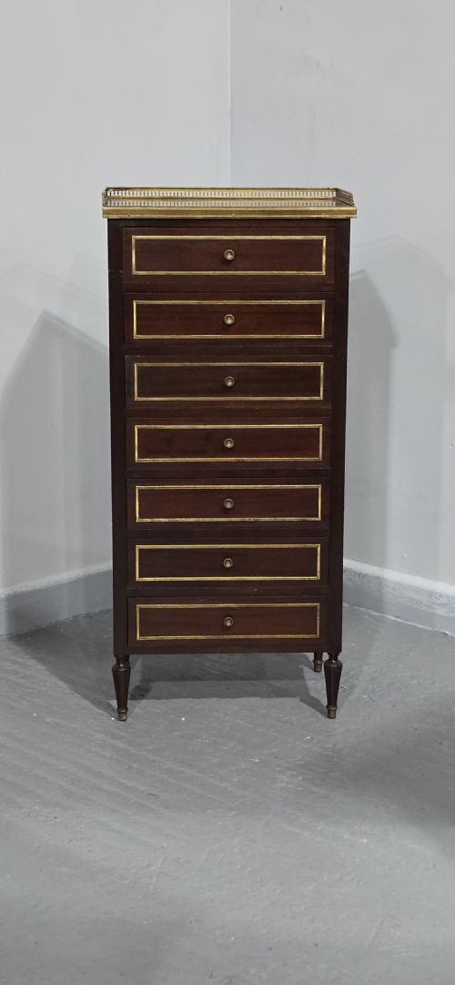 fig 1. A tall, narrow dark mahogany chest of seven drawers with brass inlay banding, single knob handles, a brass gallery top, and turned tapered legs.