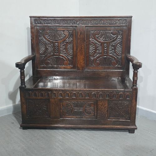 fig 1. A dark carved oak monks bench featuring relief-carved panel work on the backrest and base, complete with curved armrests and a hinged storage seat.