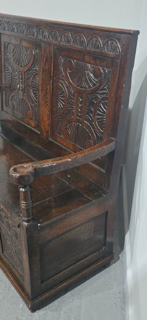 A dark carved oak monks bench featuring relief-carved panel work on the backrest and base, complete with curved armrests and a hinged storage seat. View VIII