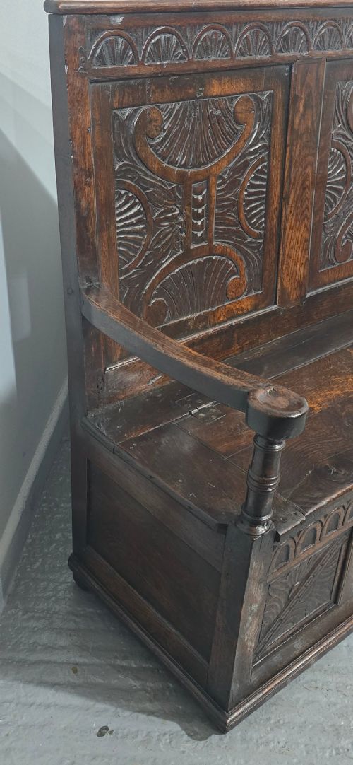 A dark carved oak monks bench featuring relief-carved panel work on the backrest and base, complete with curved armrests and a hinged storage seat. View IX