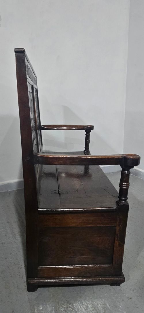 A dark carved oak monks bench featuring relief-carved panel work on the backrest and base, complete with curved armrests and a hinged storage seat. View XIII