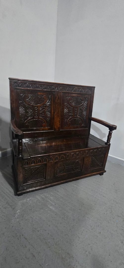 A dark carved oak monks bench featuring relief-carved panel work on the backrest and base, complete with curved armrests and a hinged storage seat. View XIV