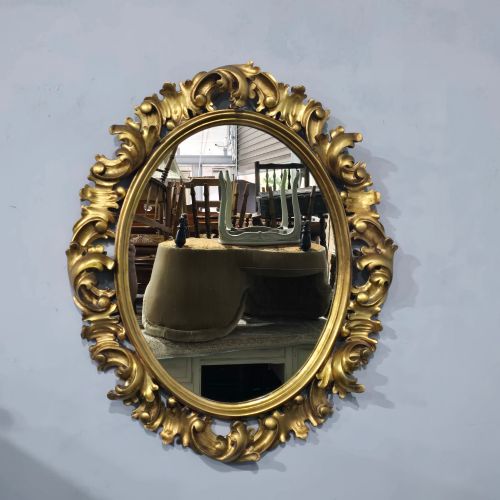 fig 1. An oval wall mirror enclosed in a deeply carved gold giltwood frame featuring ornate acanthus leaf and scrolling foliage details.