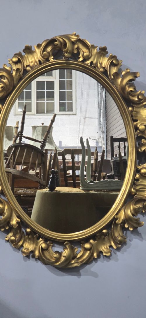 An oval wall mirror enclosed in a deeply carved gold giltwood frame featuring ornate acanthus leaf and scrolling foliage details. View IV