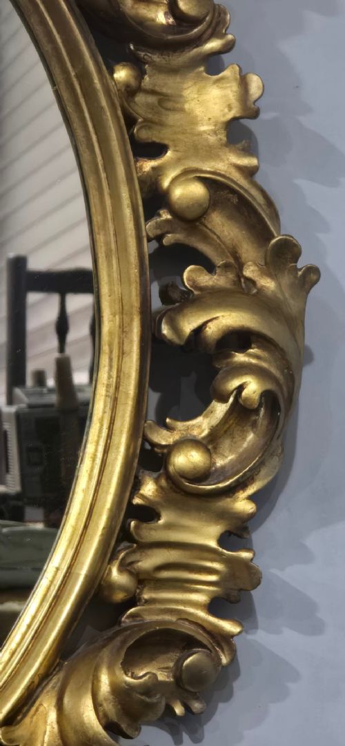 An oval wall mirror enclosed in a deeply carved gold giltwood frame featuring ornate acanthus leaf and scrolling foliage details. View VI