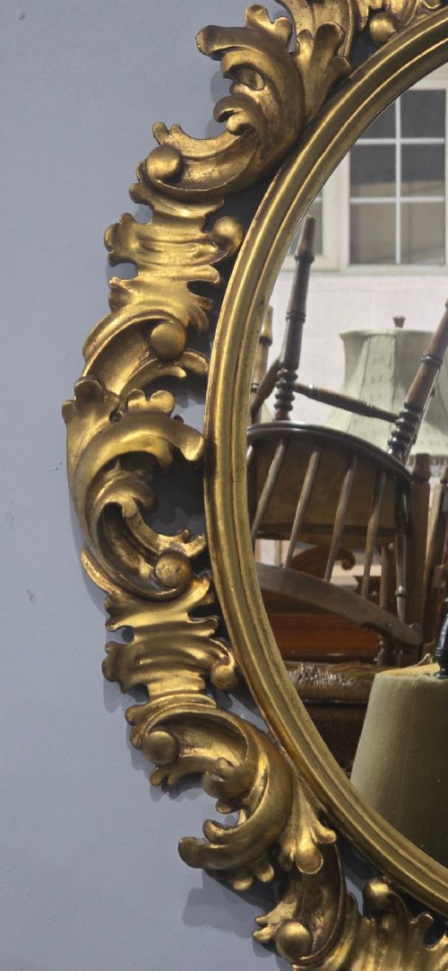 An oval wall mirror enclosed in a deeply carved gold giltwood frame featuring ornate acanthus leaf and scrolling foliage details. View IX