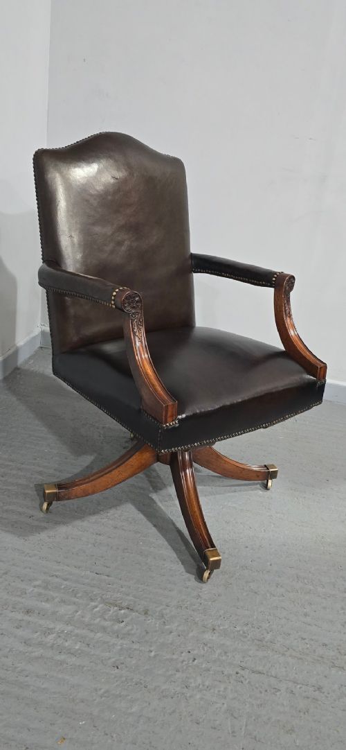 fig 1. A dark brown leather upholstered swivel desk chair featuring carved mahogany armrests, brass studding, and splayed legs with brass castors.