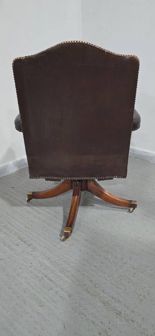 A dark brown leather upholstered swivel desk chair featuring carved mahogany armrests, brass studding, and splayed legs with brass castors. View X