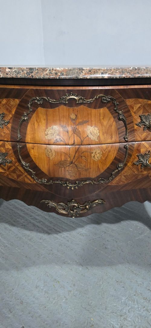 A French Louis XV style bombe commode featuring detailed floral marquetry inlay, bronze mounts, curved legs, and a brown marble top. View IV