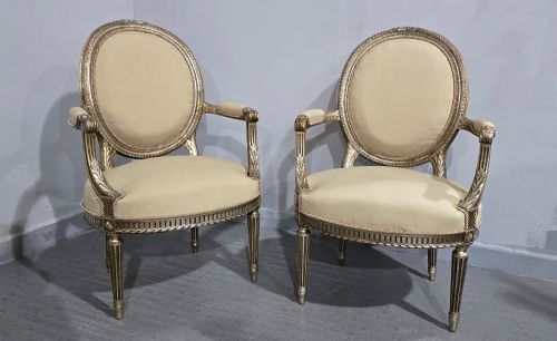 fig 1. A pair of French Louis XVI style open armchairs featuring carved giltwood frames with oval medallion backs, fluted legs, and neutral cream upholstery.