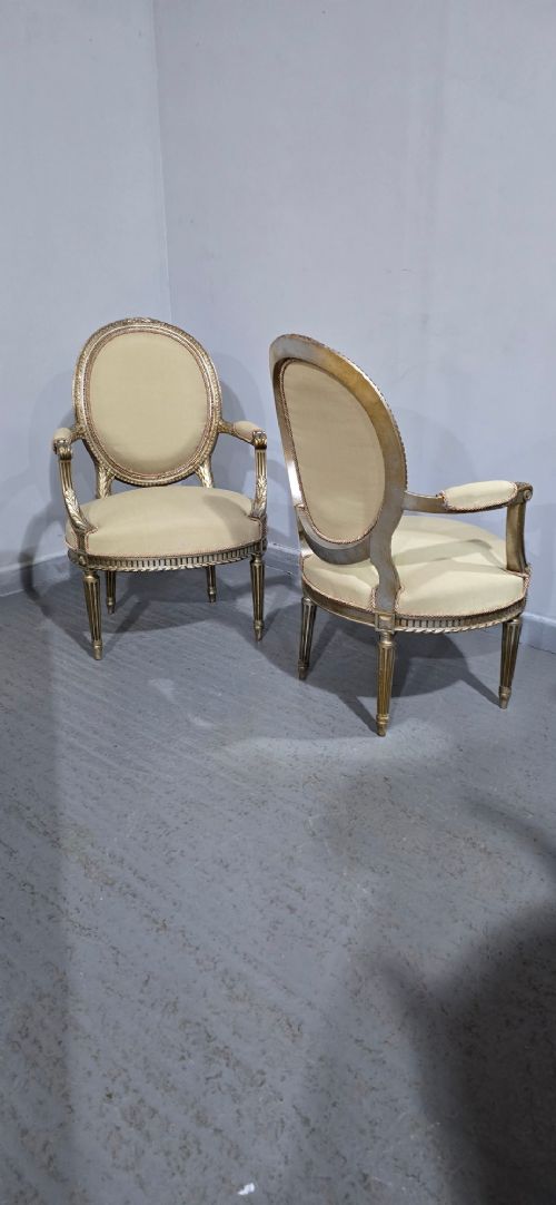 A pair of French Louis XVI style open armchairs featuring carved giltwood frames with oval medallion backs, fluted legs, and neutral cream upholstery. View IX