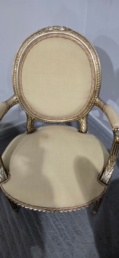 A pair of French Louis XVI style open armchairs featuring carved giltwood frames with oval medallion backs, fluted legs, and neutral cream upholstery. View X