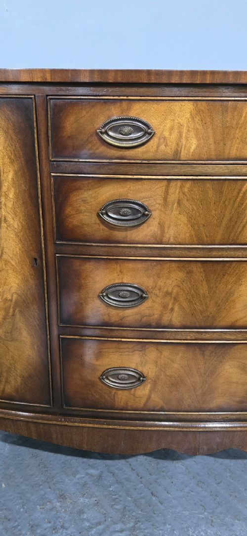 A bow-front mahogany sideboard featuring four central drawers flanked by two side cupboard doors with oval brass handles and flame mahogany grain patterns. View III