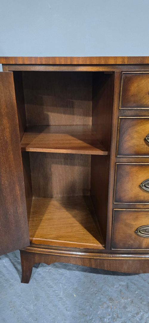 A bow-front mahogany sideboard featuring four central drawers flanked by two side cupboard doors with oval brass handles and flame mahogany grain patterns. View XI