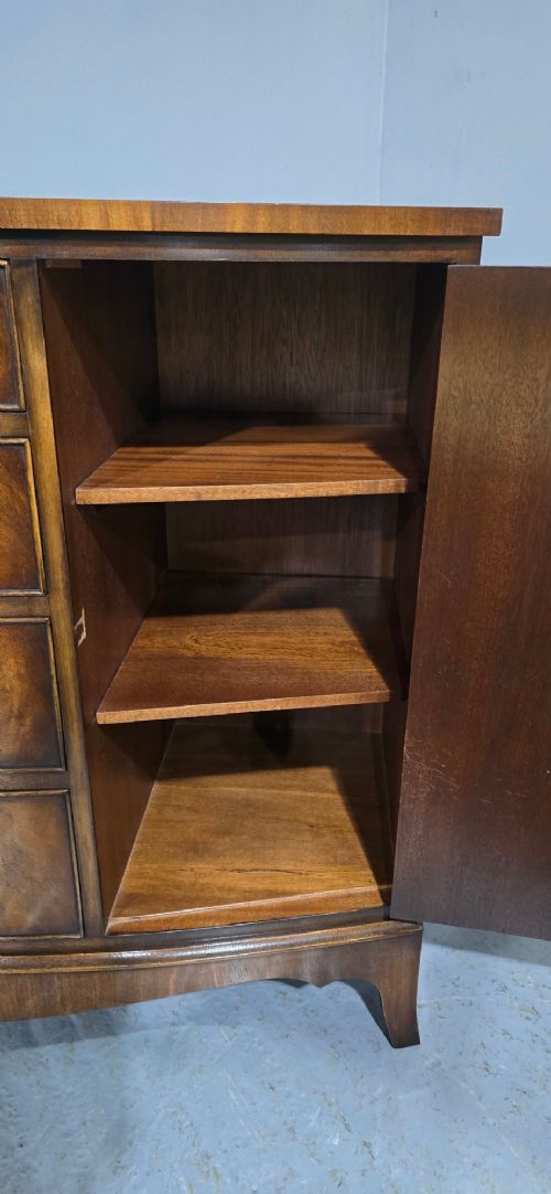 A bow-front mahogany sideboard featuring four central drawers flanked by two side cupboard doors with oval brass handles and flame mahogany grain patterns. View XII