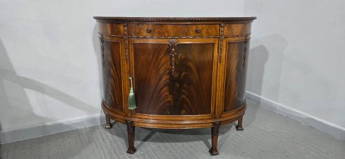 fig 1. A semi-circular mahogany demi-lune side cabinet featuring flame mahogany door panels, carved details, a single upper drawer, and tapered square feet.