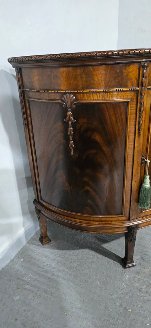 A semi-circular mahogany demi-lune side cabinet featuring flame mahogany door panels, carved details, a single upper drawer, and tapered square feet. View IV