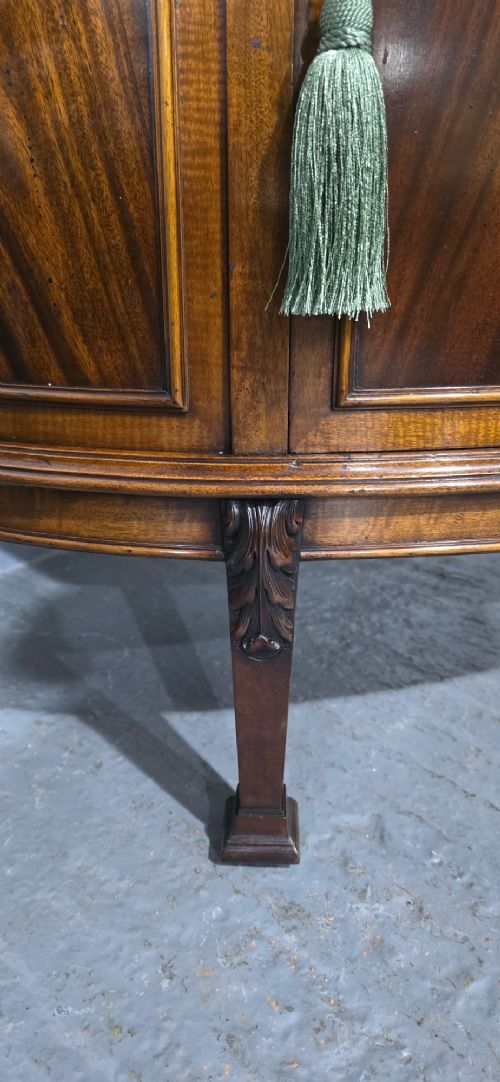 A semi-circular mahogany demi-lune side cabinet featuring flame mahogany door panels, carved details, a single upper drawer, and tapered square feet. View V