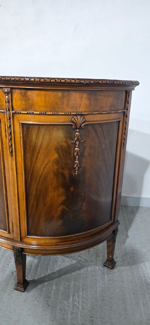 A semi-circular mahogany demi-lune side cabinet featuring flame mahogany door panels, carved details, a single upper drawer, and tapered square feet. View VIII