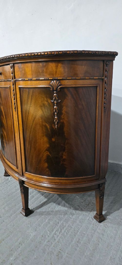 A semi-circular mahogany demi-lune side cabinet featuring flame mahogany door panels, carved details, a single upper drawer, and tapered square feet. View XIII