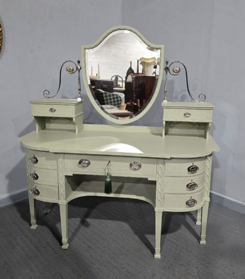 fig 1. A sage green painted dressing table featuring an integrated central shield mirror, side trinket drawers, metal scroll sconces, and tapered legs.