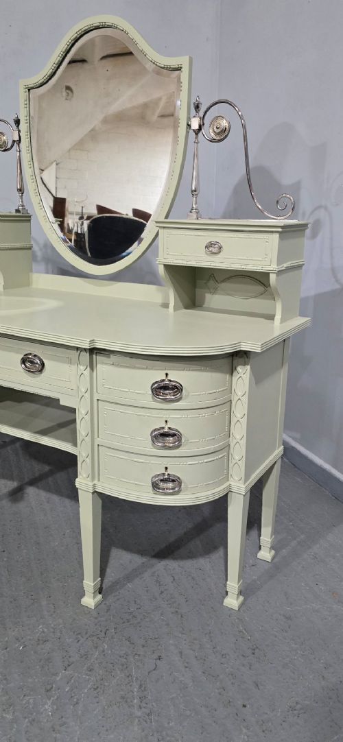 A sage green painted dressing table featuring an integrated central shield mirror, side trinket drawers, metal scroll sconces, and tapered legs. View IV