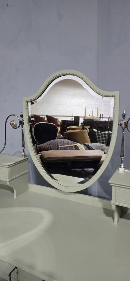 A sage green painted dressing table featuring an integrated central shield mirror, side trinket drawers, metal scroll sconces, and tapered legs. View V