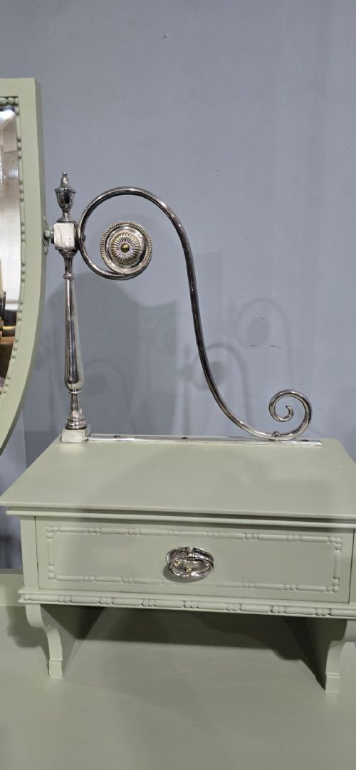 A sage green painted dressing table featuring an integrated central shield mirror, side trinket drawers, metal scroll sconces, and tapered legs. View VI