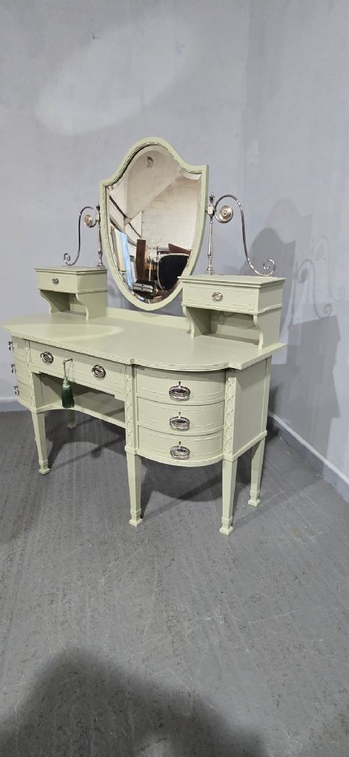 A sage green painted dressing table featuring an integrated central shield mirror, side trinket drawers, metal scroll sconces, and tapered legs. View X
