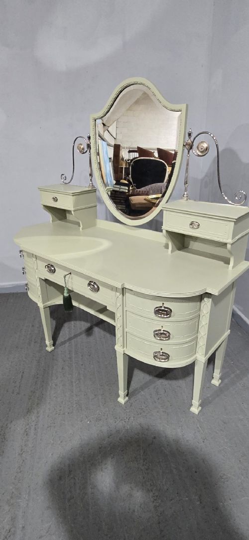 A sage green painted dressing table featuring an integrated central shield mirror, side trinket drawers, metal scroll sconces, and tapered legs. View XIV