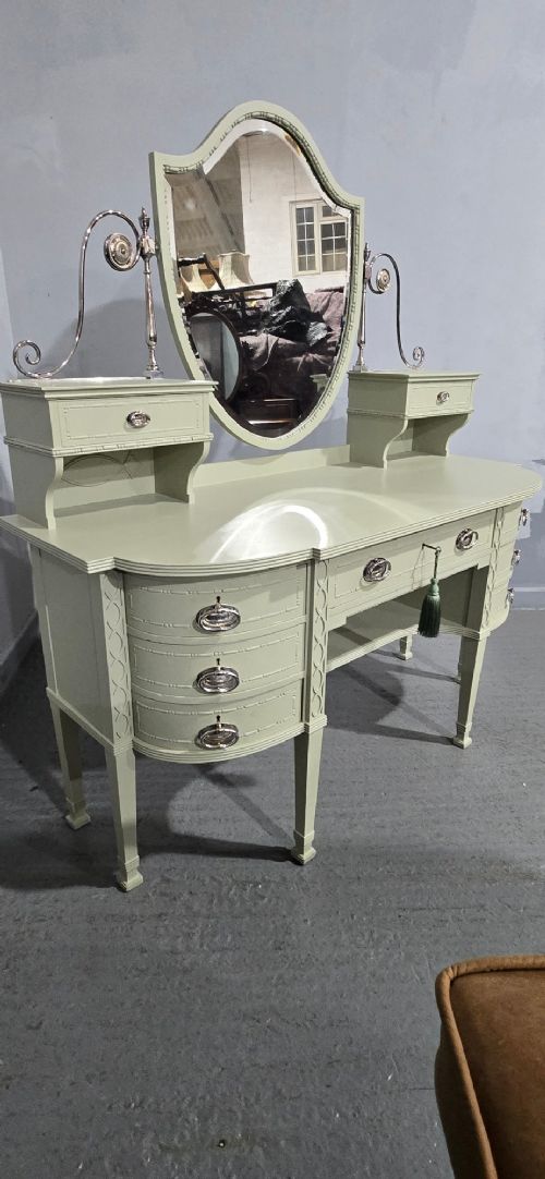 A sage green painted dressing table featuring an integrated central shield mirror, side trinket drawers, metal scroll sconces, and tapered legs. View XV