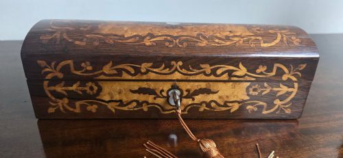 fig 1. An antique dark wood rectangular glove box featuring foliate marquetry inlay, a curved lid, a key with a silk tassel, and a mother-of-pearl escutcheon.