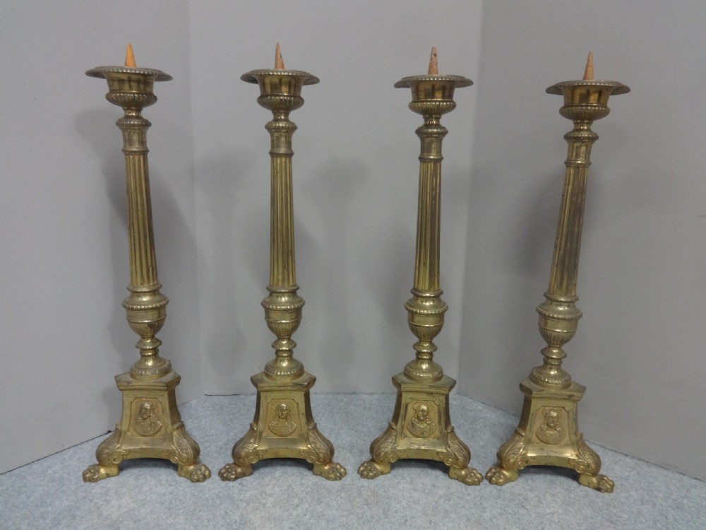 A set of four tall French brass church candlesticks featuring fluted columns, tri-form bases with relief portraits, and claw feet.