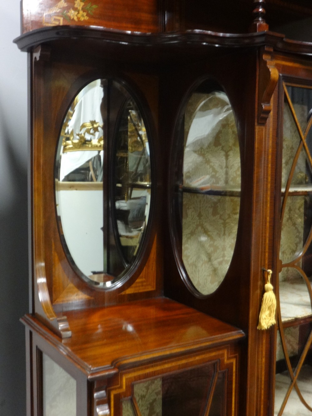 An Edwardian mahogany display cabinet featuring a broken swan-neck pediment, a central glazed door, oval mirrors, and satinwood inlay. View VI