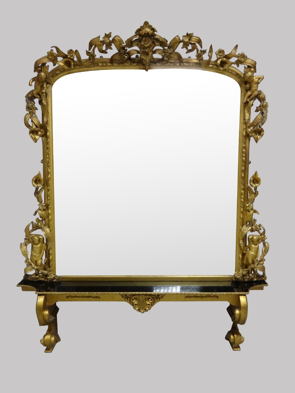 An ornate Victorian giltwood overmantel mirror featuring detailed floral cresting, cherub figures, and a black-topped console shelf supported by scroll brackets.