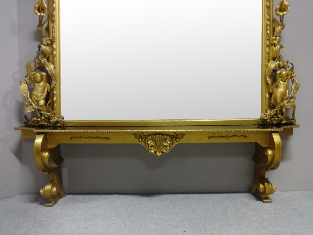 An ornate Victorian giltwood overmantel mirror featuring detailed floral cresting, cherub figures, and a black-topped console shelf supported by scroll brackets. View II