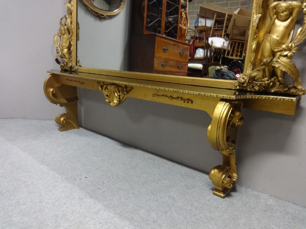 An ornate Victorian giltwood overmantel mirror featuring detailed floral cresting, cherub figures, and a black-topped console shelf supported by scroll brackets. View III