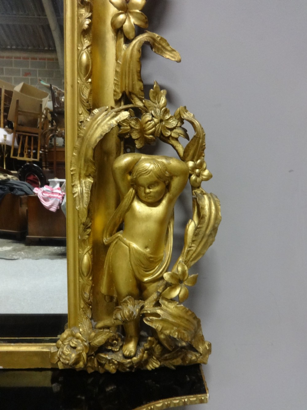 An ornate Victorian giltwood overmantel mirror featuring detailed floral cresting, cherub figures, and a black-topped console shelf supported by scroll brackets. View IV