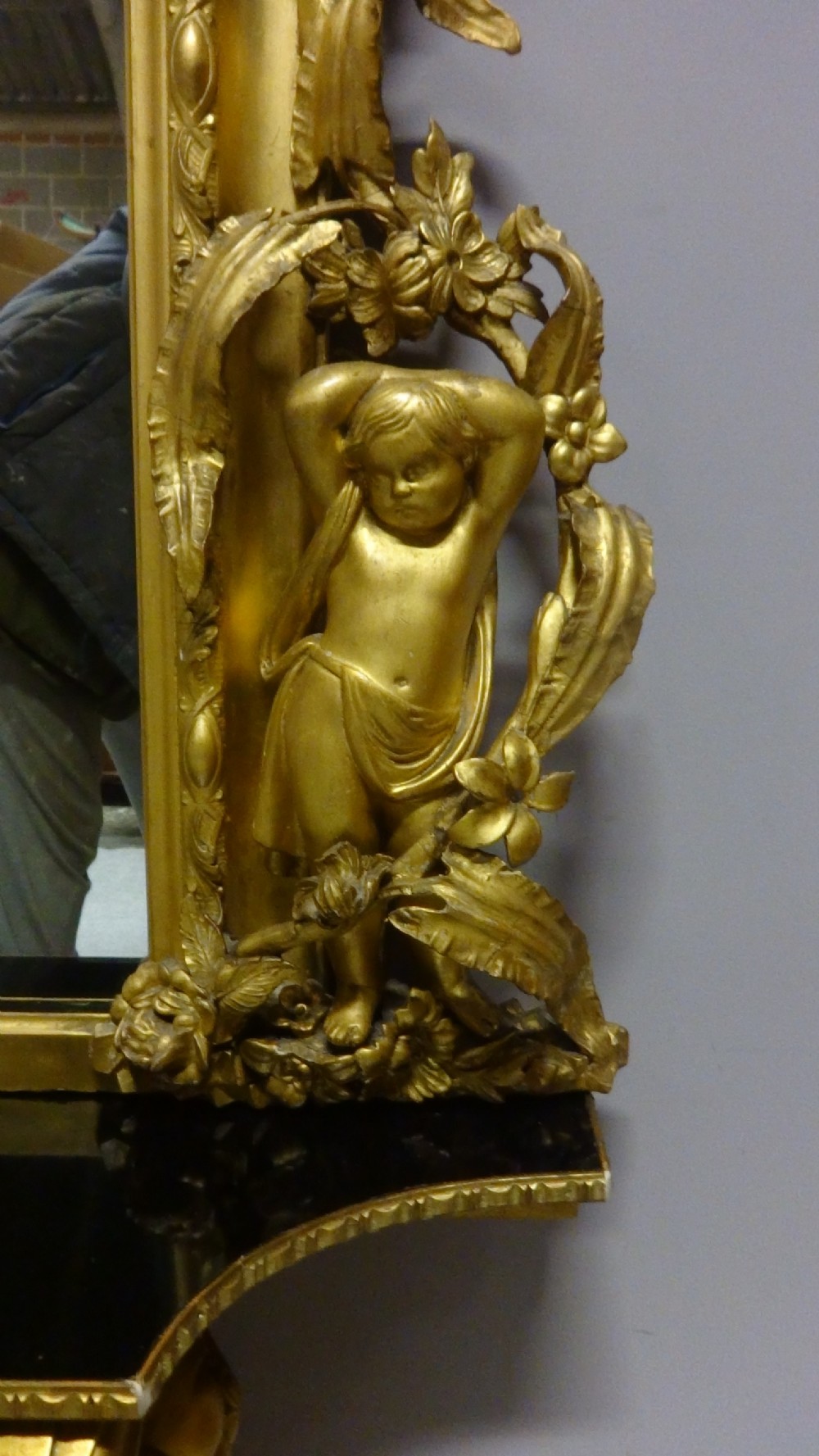 An ornate Victorian giltwood overmantel mirror featuring detailed floral cresting, cherub figures, and a black-topped console shelf supported by scroll brackets. View VI