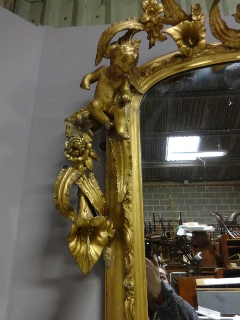 An ornate Victorian giltwood overmantel mirror featuring detailed floral cresting, cherub figures, and a black-topped console shelf supported by scroll brackets. View VII