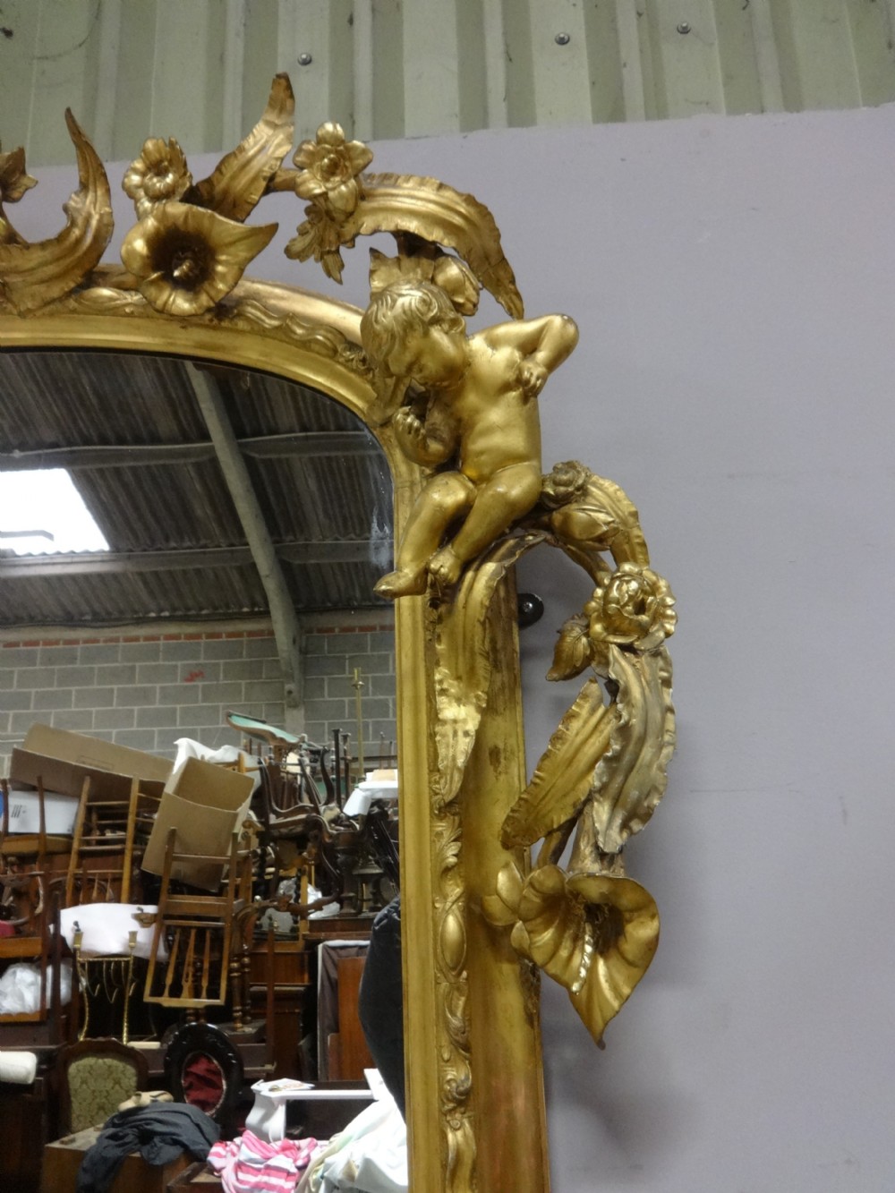 An ornate Victorian giltwood overmantel mirror featuring detailed floral cresting, cherub figures, and a black-topped console shelf supported by scroll brackets. View IX