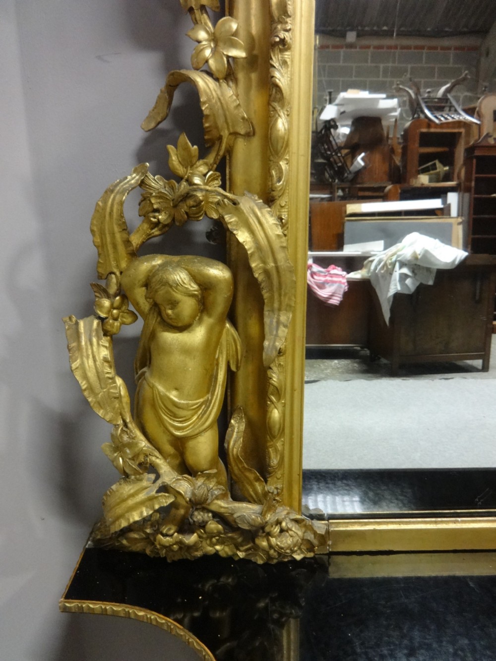 An ornate Victorian giltwood overmantel mirror featuring detailed floral cresting, cherub figures, and a black-topped console shelf supported by scroll brackets. View XI