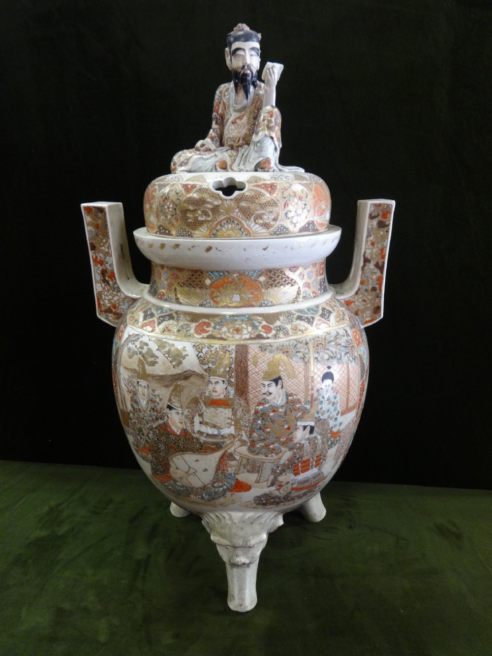 An antique Japanese Satsuma ceramic koro incense burner featuring tripodic legs, side handles, hand-painted figures, gold embellishments, and a seated figure finial on the lid.